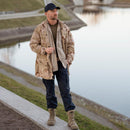 Man wearing a khaki military-style tactical shirt with black pants and boots standing by a canal outdoors