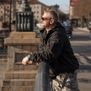 Man wearing black MIL-TEC fleece-lined military jacket with hood leaning on railing outdoors in urban setting