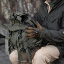 Man wearing tactical gloves holding an olive green Austrian military backpack with detachable top cover outdoors