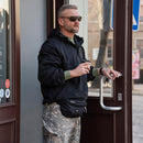 Man wearing a black MIL-TEC fleece-lined hooded military jacket with camouflage pants, holding a coffee cup at a door.