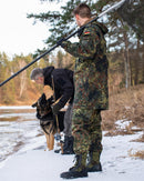 Man in German camouflage uniform wearing black and OD green Bates side zip military boots in snowy outdoor setting.