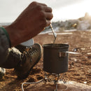 Person stirring food in a camping pot on a Primus Express Spider II tripod stove outdoors at sunset