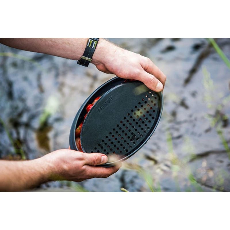 Hands holding a Primus Frigg W Piezo camping mess kit plate with food outdoors near water background