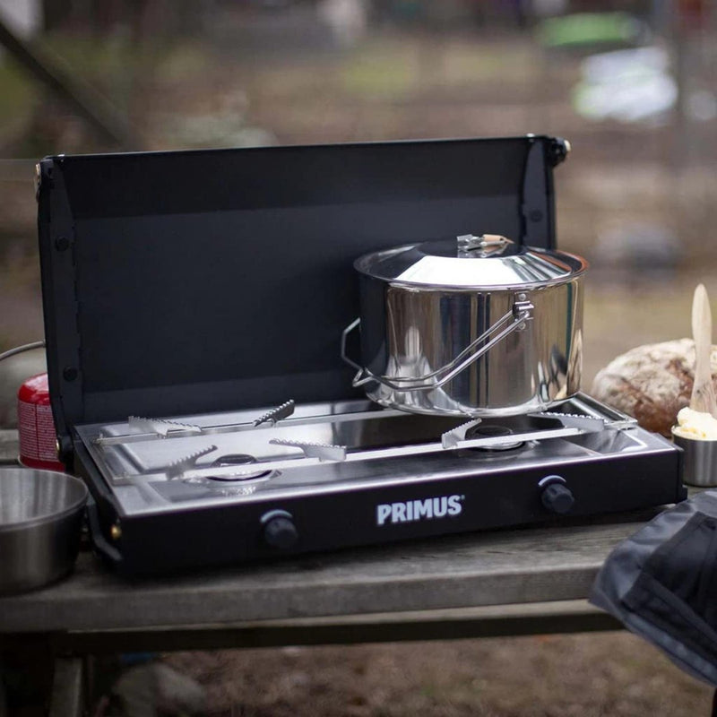 Primus Kinjia dual burner camping stove with stainless steel pot cooking outdoors on a wooden table