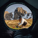 Woman using Primus PrimeTech outdoor camping cooking set with mountains in background viewed from inside tent