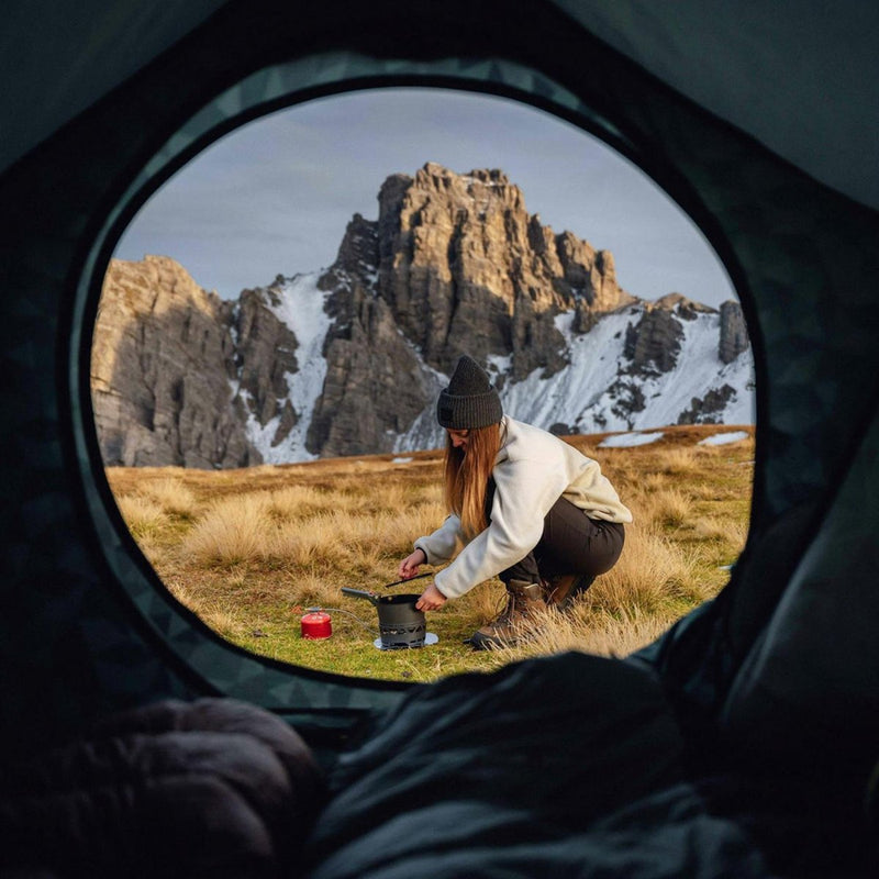 Woman using Primus PrimeTech outdoor camping cooking set with mountains in background viewed from inside tent
